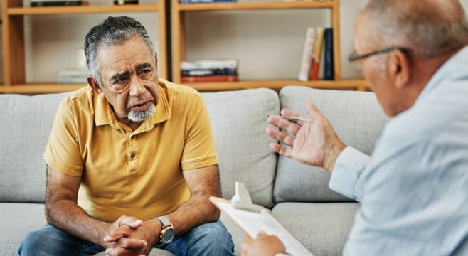 Older man listening as a therapist gestures and takes notes during a counselling session